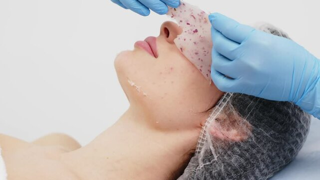 Close-up of a cosmetologist&rsquo;s hands applying an alginate mask with rose petals to a young woman&rsquo;s face in a professional beauty salon. Concept of skincare and relaxation in spa treatments.