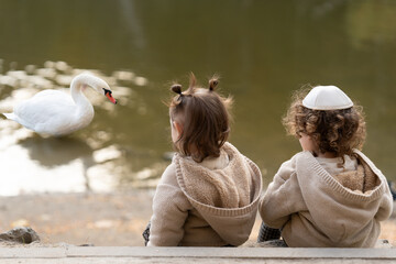 Children observe a swan by the water in a serene park setting during a calm afternoon. Small boy...