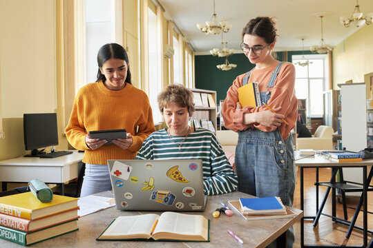 Three students engaging in collaborative study session in a spacious room with books, laptops, and tablets. Diverse group concentrating on assignments while interacting with each other