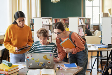 Three students gathered around laptop working on a group project in a library. Shelves of books and...