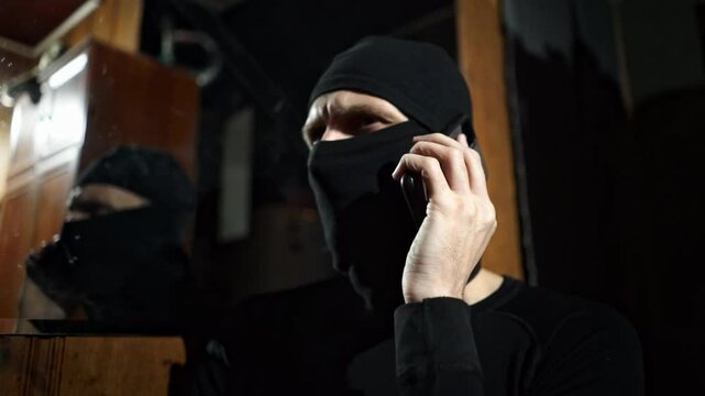 A man wearing a black mask covering most of his face is seen talking on a cell phone, with his reflection visible in a mirror. The image suggests anonymity, secrecy, or perhaps illicit activity