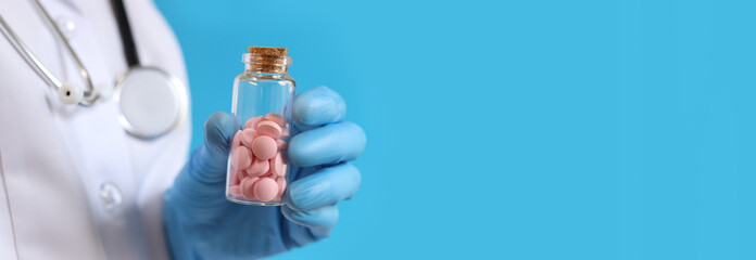 Fototapeta premium Pink pills in a glass bottle. Vitamins in a nurse or doctor's hand on a blue background. Vitamins and dietary supplements close-up. Doctor in medical gloves holds a jar of vitamins. Medicine