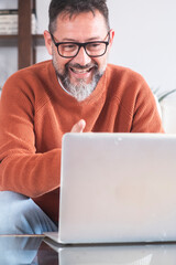 Middle-aged man wearing glasses and casual clothing, seated comfortably on a sofa at home while working remotely on a laptop, seamlessly combining freelance tasks with personal online leisure