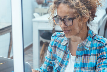 Middle-aged woman sitting at home near a window with curly hair and glasses, using her laptop for online business, remote freelance work, or browsing the internet, enjoying her professional