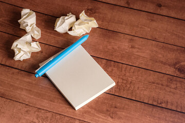 Blank notepad with blue pen on a wooden table surrounded by crumpled paper balls. Creative workspace for brainstorming and idea generation.