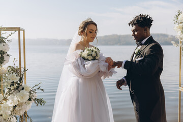 Groom placing wedding ring on bride's finger during interracial wedding ceremony by the lake