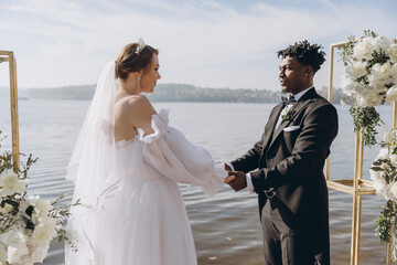 Interracial couple holding hands during wedding ceremony by the lake