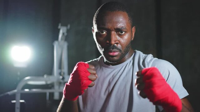Boxer in gym. Aggressive African man fighter resting after training boxing ready for fight looking at camera. Strong sweated man training punches looking concentrated straight preparing for sparring