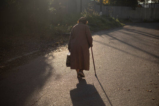 Senior man walking on road in sunlight. Retired woman with walking stick in autumn. An old woman in a coat.