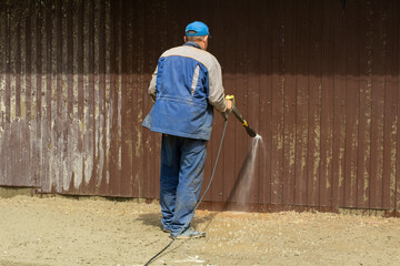 Worker washes dirt off fence. Water hose. Cleaning area. Sand laundering from steel fence.