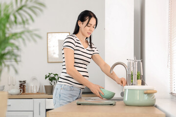 Young pregnant woman with bowl turning on tap in kitchen