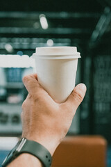 Mão segurando copo descartável de café em ambiente urbano moderno com iluminação suave. Fotografia minimalista, destacando estilo de vida contemporâneo, pausa, conforto e momentos de café em movimento