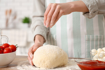 Woman sprinkling raw dough with flour for pizza near table in kitchen