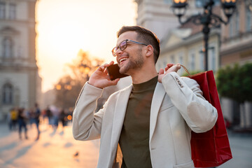 Caucasian man in white coat speaks on smartphone while carrying red shopping bag through historic...