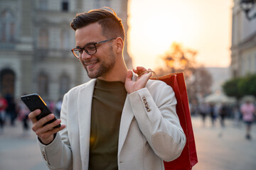 Caucasian male in white blazer checks smartphone while carrying red shopping bag on city street...