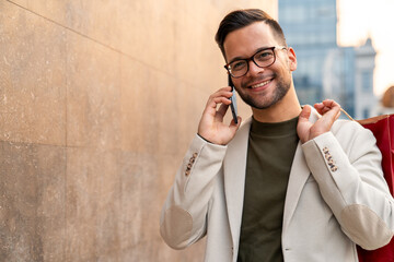 Caucasian male professional in beige blazer and olive shirt smiles while talking on smartphone and carrying shopping bag along city wall.