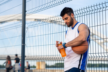 Athletic male in white basketball jersey hydrates with orange sports drink while checking fitness tracker beside outdoor court fence.