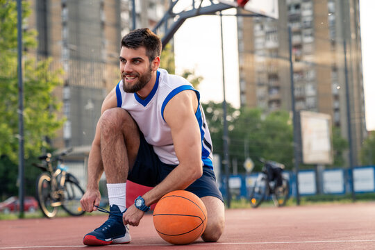 Athletic male in basketball uniform prepares for game on outdoor court surrounded by urban apartment buildings and trees.