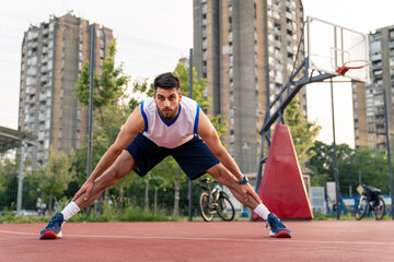 Athletic male in basketball jersey performs stretching routine on outdoor court surrounded by...