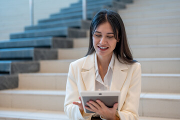 woman in white blazer smiles while using tablet on stairs, showcasing professional and relaxed atmosphere. Her engagement with technology reflects modern work culture