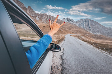 Woman making rock and roll hand gesture from car window while enjoying road trip in the mountains