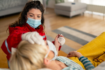 Female emergency responder in red uniform and protective mask provides medical care to elderly...