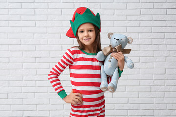 Cute little girl in elf costume with plush toy near white brick wall. Christmas celebration