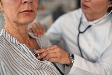 Female physician in white coat performs thorough medical examination using stethoscope on senior patient wearing striped shirt during residential healthcare visit.