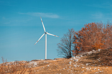 White wind turbine generating renewable energy on a mountain with autumnal trees and blue sky