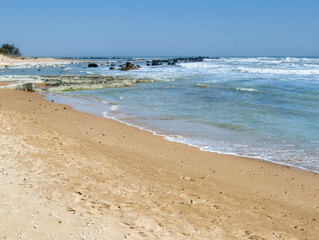 Golden sandy beach with small rocks and clear turquoise waters, gently lapping under a bright blue sky near Scala dei Turchi. Sicily, Italy.