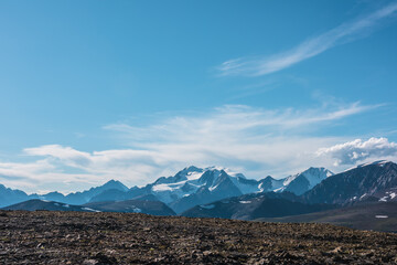 Big snow-capped mountain top far away in sunny day. Scenic alpine top view from sunlit high stony plain of pass to large rocky snowy mountain peak with glacier in bright sun under clouds in blue sky.