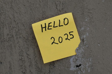 Una toma de cerca de una nota adhesiva con la palabra hola y el numero 2025 escrita a mano  pegada a una pared © Elika R