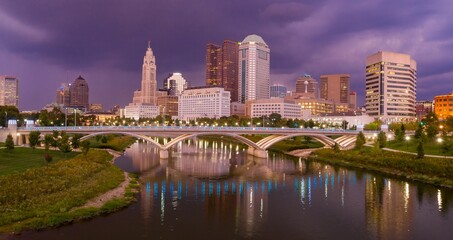 Naklejka premium Evening cityscape of downtown Columbus, Ohio, reflected in the Scioto River. Modern architecture, illuminated bridge, and tranquil water. DOWNTOWN, COLUMBUS, OHIO, UNITED STATES
