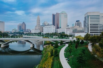 Runner on paved path, city skyline reflected in river, urban park. EAST BANK PARK, COLUMBUS, OHIO,...