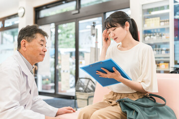 Fototapeta premium 病院・薬局の待合室で問診票を書く女性と相談に乗る薬剤師 