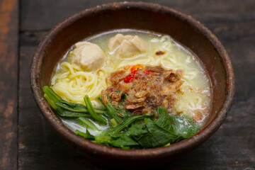 Noodle soup with vegetables, meatballs, and fried shallots, served in a clay bowl placed on a dark wooden table. A hearty and traditional dish perfect for showcasing Indonesian comfort food.