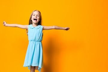 Joyful little girl in a blue dress standing with arms outstretched against a vibrant orange...