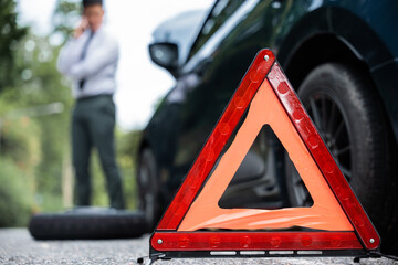 Side view of a businessman sitting on a warning triangle beside his broken car. Repairing the car...