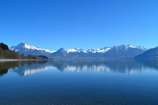 A panoramic view of an Alpine lake surrounded by majestic snow-capped mountains in Switzerland, with a clear blue sky reflecting on the tranquil water.