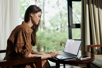 Young woman working on a laptop at a table in a cozy setting with natural light streaming in Focus...