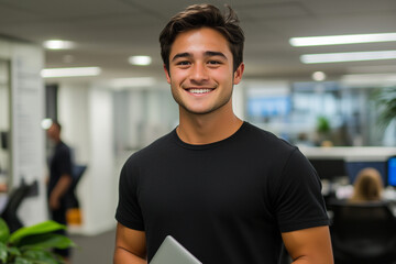 Fototapeta premium Smiling Young Professional Holding a Notebook in a Bright, Modern Office with an Open-Plan Layout and Lush Green Plants Happy Businessman in a Black T-Shirt, Standing in a Contemporary Office Setting 