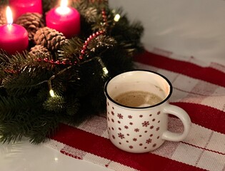 Top view of a white coffee mug on table with green wreath and burning candles 