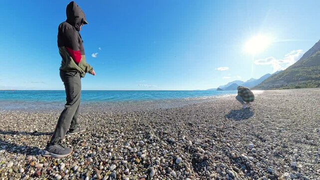 A man and woman throwing stones into the sea on a cool, bright day, standing on a pebbled beach with turquoise water, green cliffs, and a clear blue sky.
