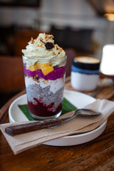 A healthy breakfast with a chia pudding and fruit, jam, granola and cream in a tall glass cup with a spoon on a plate. Vegan dessert. Vertical shot