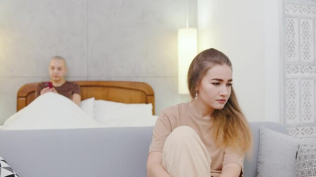 A sad lesbian woman sits on the couch, lost in thought, while her bald girlfriend lies in bed in the background, indifferently using her smartphone.