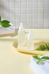 Mockup of dropper bottles placed on a wooden tray, next to some towels, green tea leaves, and fern fronds, in a bathroom setting. Advertising photos for serum products.