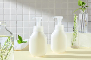 In the center are two white pump bottles no label, surrounded by some glass vases containing a green tea leaves and fern branches, place on a yellow surface, in a bathroom backdrop. Advertising photo.