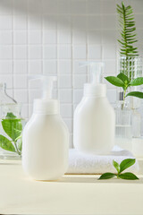 Two white pump bottles without labels, displayed on a towel on a yellow surface, are surrounded by green tea leaves and fern fronds in glass vases. Minimal bathroom setting for body care products ads.