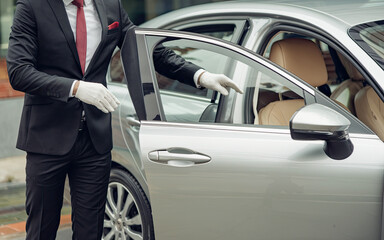 Man in Formal Black Suit, White Shirt, and Red Tie Standing Next to Silver Car, Wearing White Gloves, Opening Car Door with Beige Interior, Outdoors Setting, Street or Parking Lot
