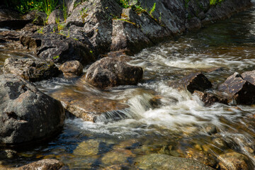 Fototapeta premium Norwegian mountain river with fast flowing clear and sparkling water and a waterfall that forms white foam on rocks surrounded by green coniferous trees. Sunny summer day.